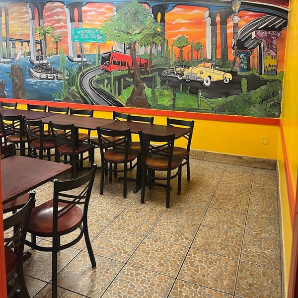 Dining area with dark tables and chairs, featuring a vibrant San Diego mural on the wall.